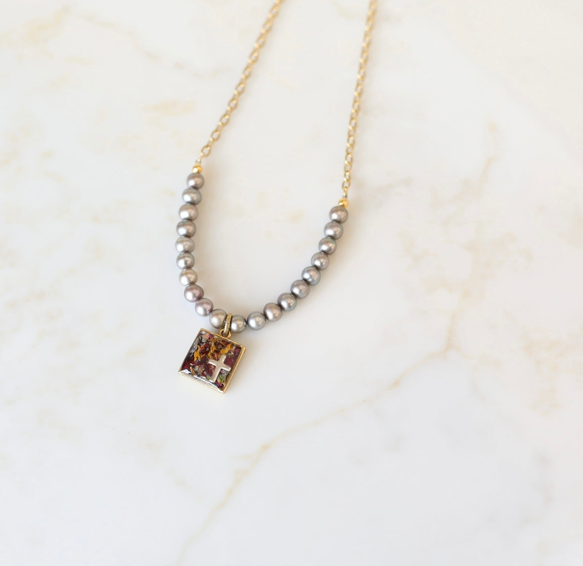 A silver-colored necklace featuring a square freshwater pearl pendant with a cross charm, surrounded by a chain of pearls on a marble surface.