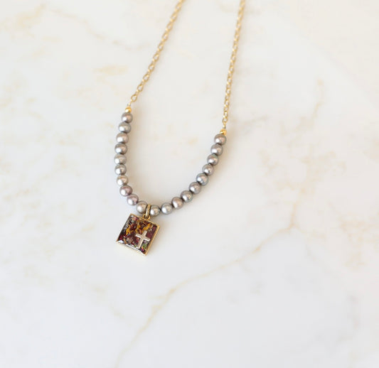 A silver-colored necklace featuring a square freshwater pearl pendant with a cross charm, surrounded by a chain of pearls on a marble surface.